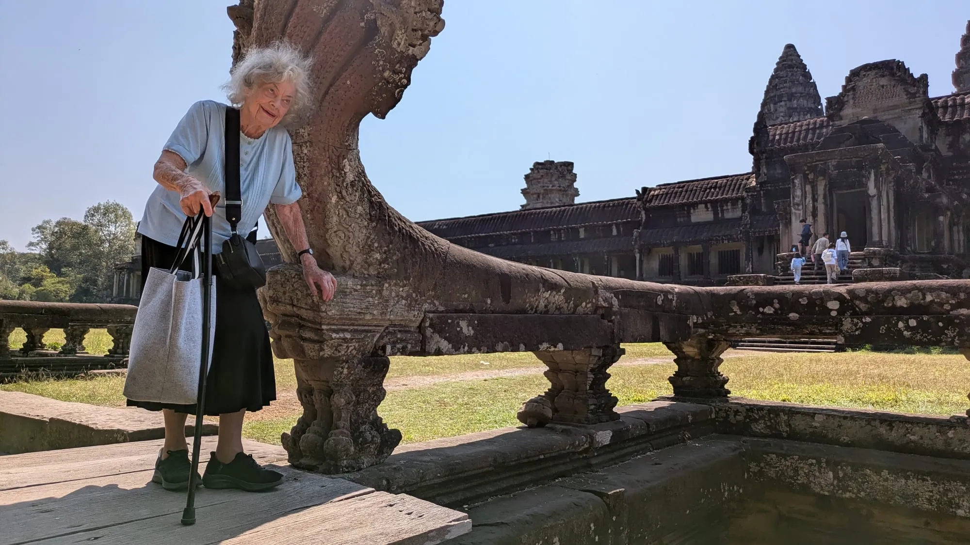 Dee leaning on her cane beside a carved stone balustrade, with the towers of Angkor Wat behind