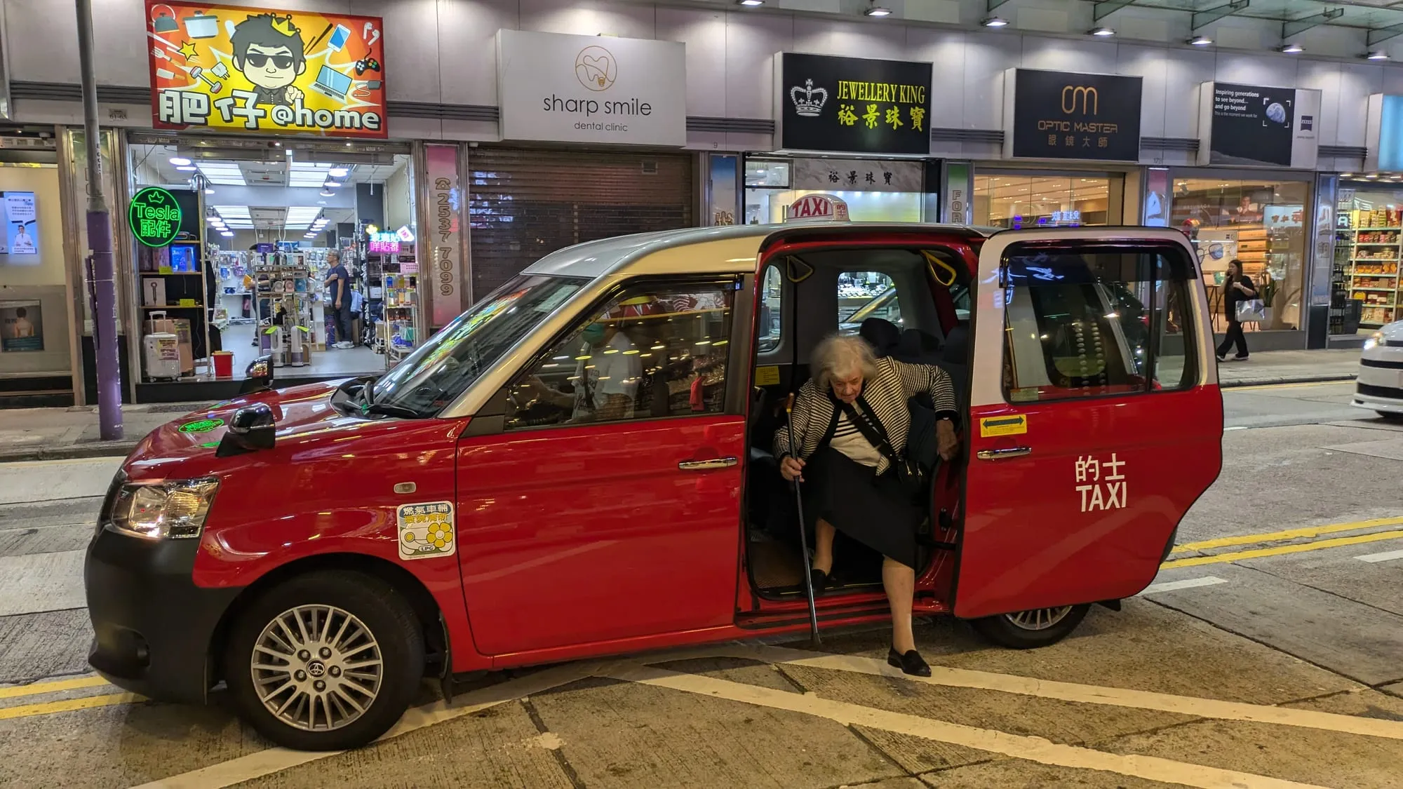Dee climbing out of a red Hong Kong taxi on a busy street at night