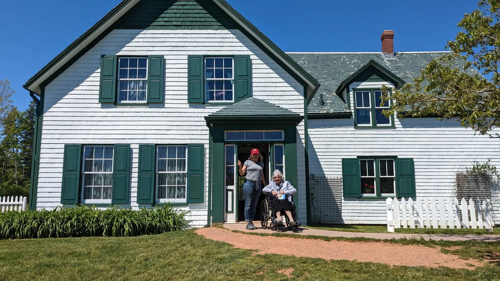 Dee in a wheelchair at the door of a white farmhouse with green shutters and gables
