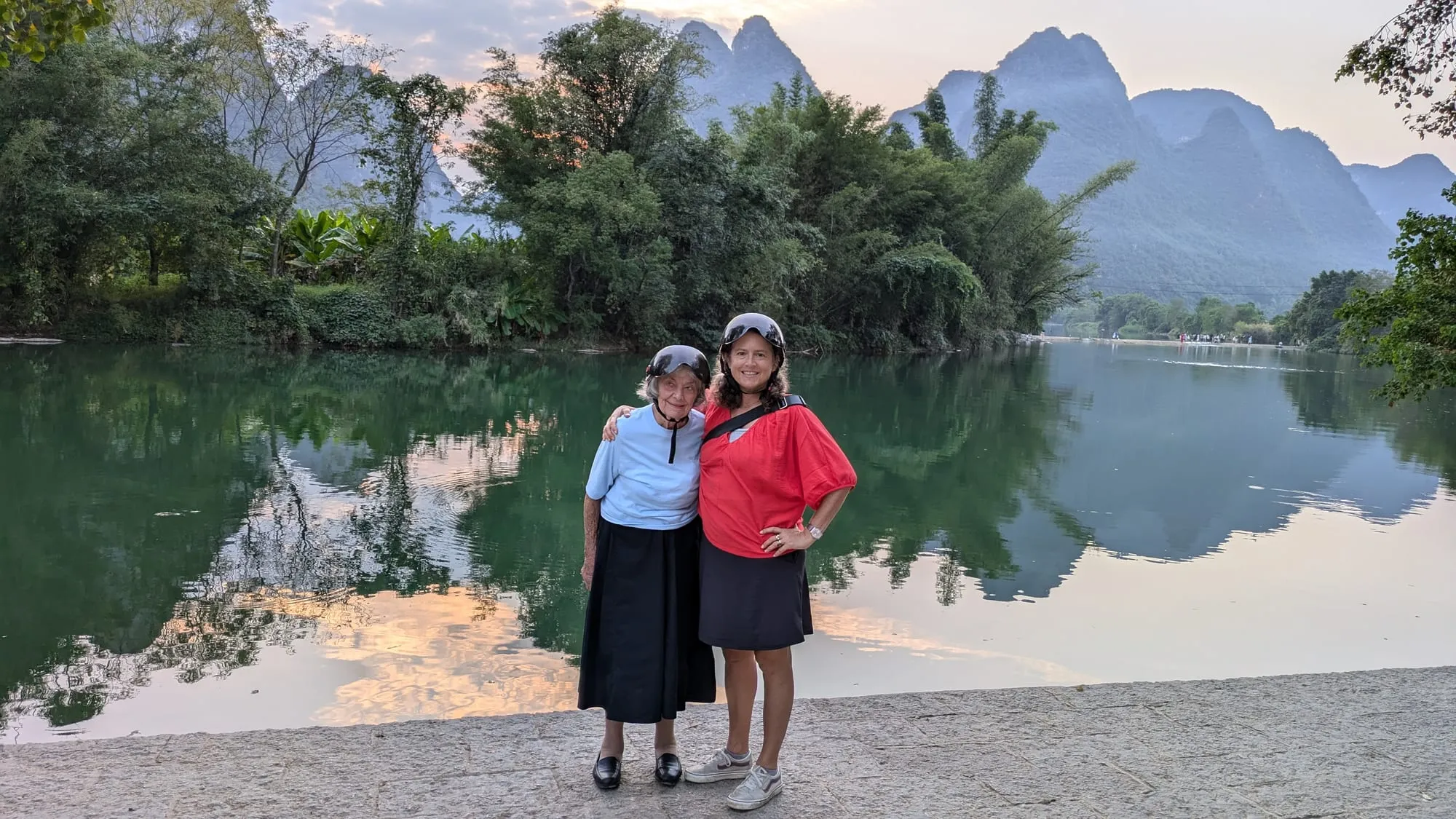 Dee and Kim by a still river reflecting jagged limestone peaks at dusk